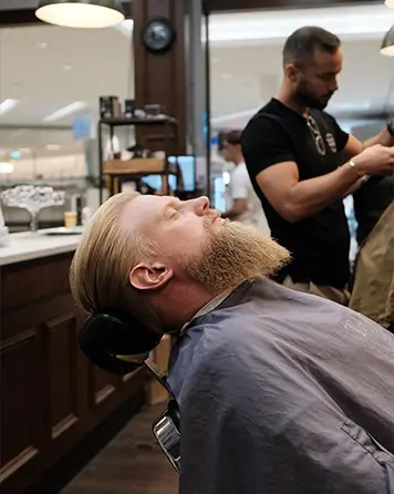 Man med långt skägg lutar bakåt i stolen under kombinationsbehandlingen herrklippning och skäggtrimning.