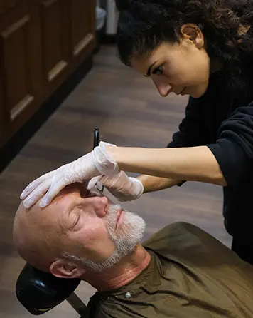 Barberare utför rakning med kniv på kund som ligger i barberarstol.